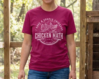 Può includere: Una t-shirt a maniche corte color magenta con una stampa grafica bianca di tre polli e il testo I HAVE A COUPLE CHICKENS CHICKEN MATH. La maglietta è abbinata a jeans blu, creando un outfit casual adatto per le attività all'aperto.