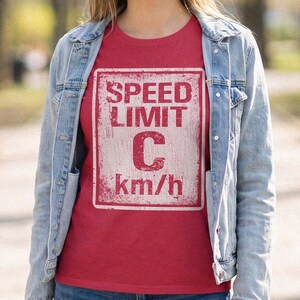 May include: A red short-sleeved t-shirt featuring a white rectangular graphic that resembles a road sign. The graphic displays the text SPEED LIMIT, a large letter C, and the text km/h, all presented with a distressed, weathered texture.