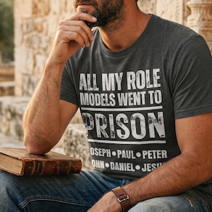May include: A dark gray t-shirt with the text "ALL MY ROLE MODELS WENT TO PRISON" in white. Below the word "PRISON" are the names "OSEPH PAUL PETER OHN DANIEL JESU". The person is wearing blue jeans and a brown leather watch.