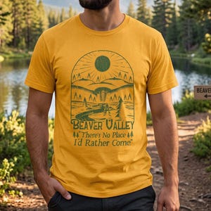 May include: A mustard yellow short-sleeve t-shirt featuring a green graphic print of a landscape with mountains, trees, and a sun. The text reads: Beaver Valley, There's No Place I'd Rather Come, with small pine tree icons on either side.