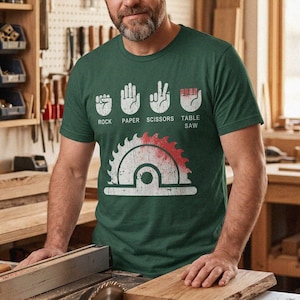 May include: A green t-shirt with a white graphic of a table saw and the words "Rock Paper Scissors Table Saw". The image shows a man in a workshop with woodworking tools and a table saw.
