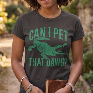 May include: A dark grey short-sleeved t-shirt featuring a green graphic of an alligator in the centre. The text "CAN I PET THAT DAWG?" is printed in green capital letters above and below the alligator illustration on the front of the shirt.
