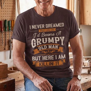 Peut inclure: Un t-shirt à manches courtes marron foncé avec le texte blanc et orange : I NEVER DREAMED THAT ONE DAY I'd Become A GRUMPY OLD MAN BUT HERE I AM KILLIN IT. Le t-shirt est porté avec un jean bleu, offrant un style décontracté et confortable.