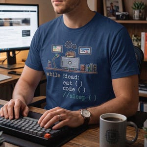 May include: A navy blue t-shirt featuring a graphic of a desk setup with a computer and the text while !dead: eat () code () //sleep (). The design includes small icons of gears, screens, and books, printed in white, blue, and green ink.