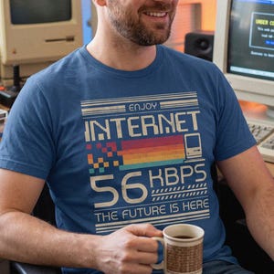 May include: A blue short-sleeved t-shirt featuring a retro-style graphic with the text "ENJOY INTERNET 56 KBPS THE FUTURE IS HERE," a pixelated rainbow bar, and a small computer icon, all printed in a distressed, vintage-inspired font and style.