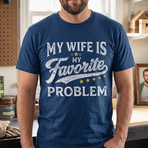 Puede incluir: Una camiseta azul marino de manga corta con texto blanco de estilo desgastado que dice MY WIFE IS MY FAVORITE PROBLEM, con la palabra favorite en fuente cursiva y cinco pequeñas estrellas amarillas situadas encima y debajo del texto.