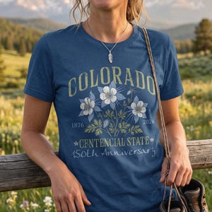 May include: A navy blue short-sleeve t-shirt featuring a graphic of white and blue columbine flowers. The text reads COLORADO, 1876, 2026, CENTENIAL STATE, and 150th Anniversary. The design is centered on the chest of the garment.