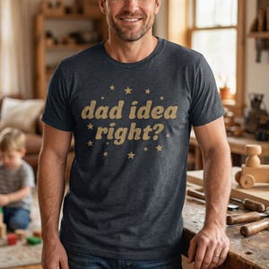 Può includere: Una t-shirt a maniche corte grigio antracite scuro con la scritta "dad idea right?" stampata in un carattere dorato dall'aspetto vissuto, circondata da piccole grafiche a forma di stella dorata. La maglia ha un girocollo e vestibilità standard.