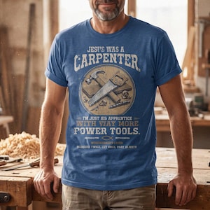Könnte beinhalten: Ein blaues kurzärmeliges T-Shirt mit einem Grafikdruck, der einen Hammer und eine Säge in einem Kreis zeigt. Der Text lautet: JESUS WAS A CARPENTER, I'M JUST HIS APPRENTICE WITH WAY MORE POWER TOOLS und MEASURE TWICE, CUT ONCE, PRAY ALWAYS.