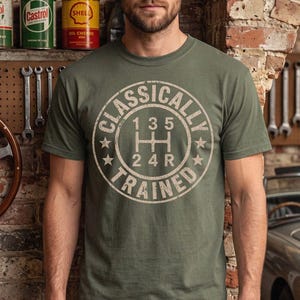 Könnte beinhalten: Ein olivgrünes Kurzarm-T-Shirt mit einer kreisförmigen Grafik auf der Brust. Das Design zeigt ein manuelles Schaltgetriebemuster mit den Zahlen 1, 3, 5, 2, 4 und R, umgeben von dem Text "CLASSICALLY TRAINED" und einigen Sternen.