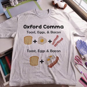 May include: A light gray short-sleeve t-shirt featuring the text Oxford Comma, Toast, Eggs, & Bacon with illustrations of toast, an egg, and bacon separated by plus signs, followed by Toast, Eggs & Bacon with illustrations of toast, eggs, and bacon.