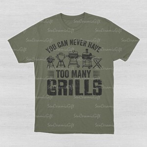 May include: An olive green short-sleeve t-shirt featuring a black graphic print. The design includes illustrations of various barbecue grills above the text "YOU CAN NEVER HAVE TOO MANY GRILLS" in a distressed, bold, block-style font.