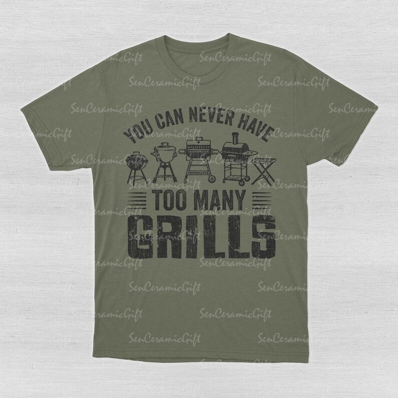 May include: An olive green short-sleeve t-shirt featuring a black graphic print. The design includes illustrations of various barbecue grills above the text "YOU CAN NEVER HAVE TOO MANY GRILLS" in a distressed, bold, block-style font.