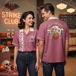 May include: A mauve short-sleeved button-down bowling shirt with a cream collar and trim. The front features a vertical bat pattern and a logo with the text Lucky Rollers and Cooper. The back displays a graphic of bowling pins and a pumpkin.