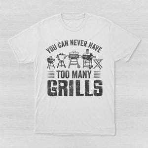May include: A white short-sleeved t-shirt featuring a black graphic print. The design displays illustrations of five different types of grills above the text "YOU CAN NEVER HAVE TOO MANY GRILLS" in a distressed, bold, sans-serif font style.