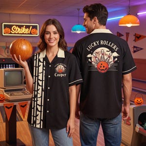 May include: A black button-down bowling shirt with a cream collar and a vertical panel featuring a black bat pattern. The front displays the text Lucky Rollers and Cooper above a bowling ball graphic. The back features a large Lucky Rollers graphic.