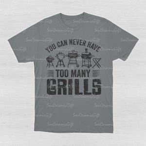May include: A grey short-sleeved t-shirt featuring a black graphic print of various barbecue grills and the text "YOU CAN NEVER HAVE TOO MANY GRILLS" in a distressed font. The design is centered on the chest of the shirt.