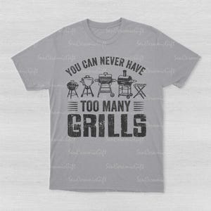 May include: A light gray short-sleeve t-shirt featuring a black graphic print. The design includes illustrations of five different types of grills above the text "YOU CAN NEVER HAVE TOO MANY GRILLS" in a bold, distressed, block-style font.