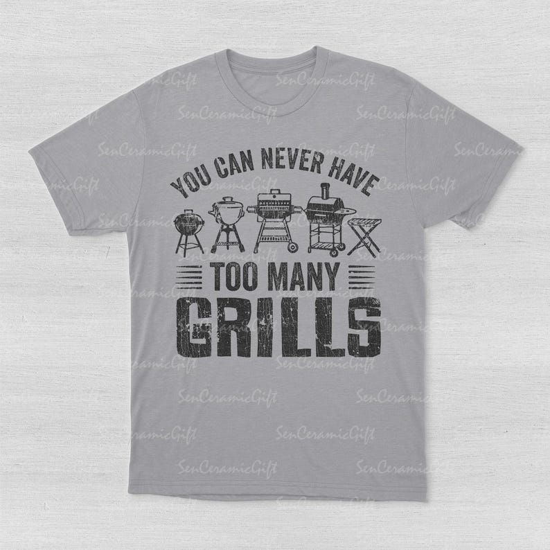 May include: A light gray short-sleeve t-shirt featuring a black graphic print. The design includes illustrations of five different types of grills above the text "YOU CAN NEVER HAVE TOO MANY GRILLS" in a bold, distressed, block-style font.