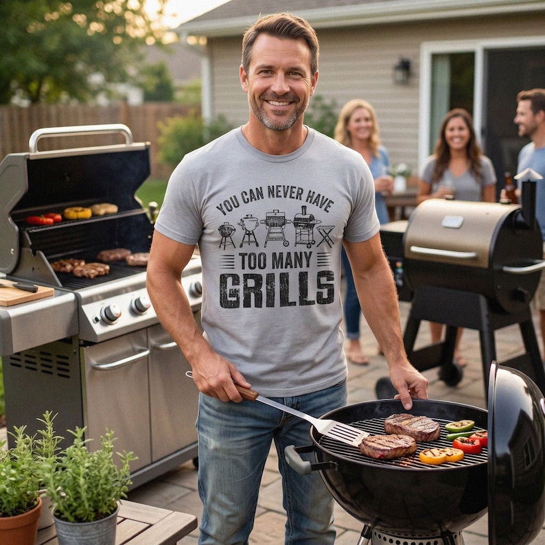 May include: A light gray short-sleeve t-shirt featuring black text that reads "YOU CAN NEVER HAVE TOO MANY GRILLS" above a graphic illustration of five different types of barbecue grills. The shirt is made of a soft cotton-blend fabric.
