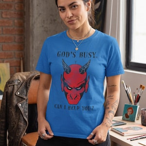 Puede incluir: Una camiseta de manga corta de color azul brillante con un gráfico de una cara de diablo roja con cuernos. El texto "GOD'S BUSY." está impreso sobre la ilustración del diablo, y el texto "CAN I HELP YOU?" está impreso debajo en negro.