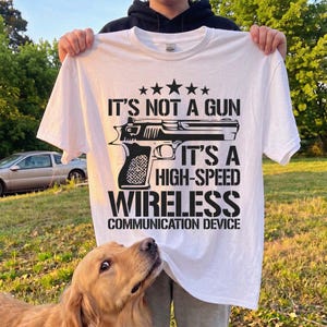 Peut inclure: Un t-shirt blanc à manches courtes avec un motif noir représentant une arme de poing et le texte : IT'S NOT A GUN IT'S A HIGH-SPEED WIRELESS COMMUNICATION DEVICE, surmonté de cinq étoiles disposées en ligne horizontale.