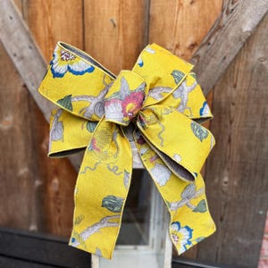 May include: A large, decorative bow with a vibrant yellow background and a floral pattern in shades of pink, blue, and gray. The bow is placed on top of a white lantern with ornate detailing, set against a rustic wooden backdrop.