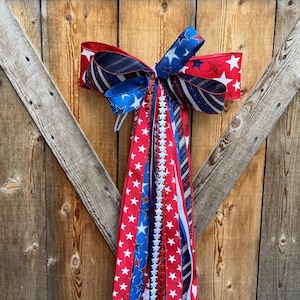 May include: A red, white, and blue bow with stars and stripes. The bow is attached to a long, flowing ribbon with stars and stripes. The bow is hanging on a wooden door.