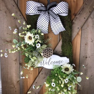May include: A welcome wreath with a black and white polka dot bow, a white sign that says "Welcome", and white flowers with green leaves.