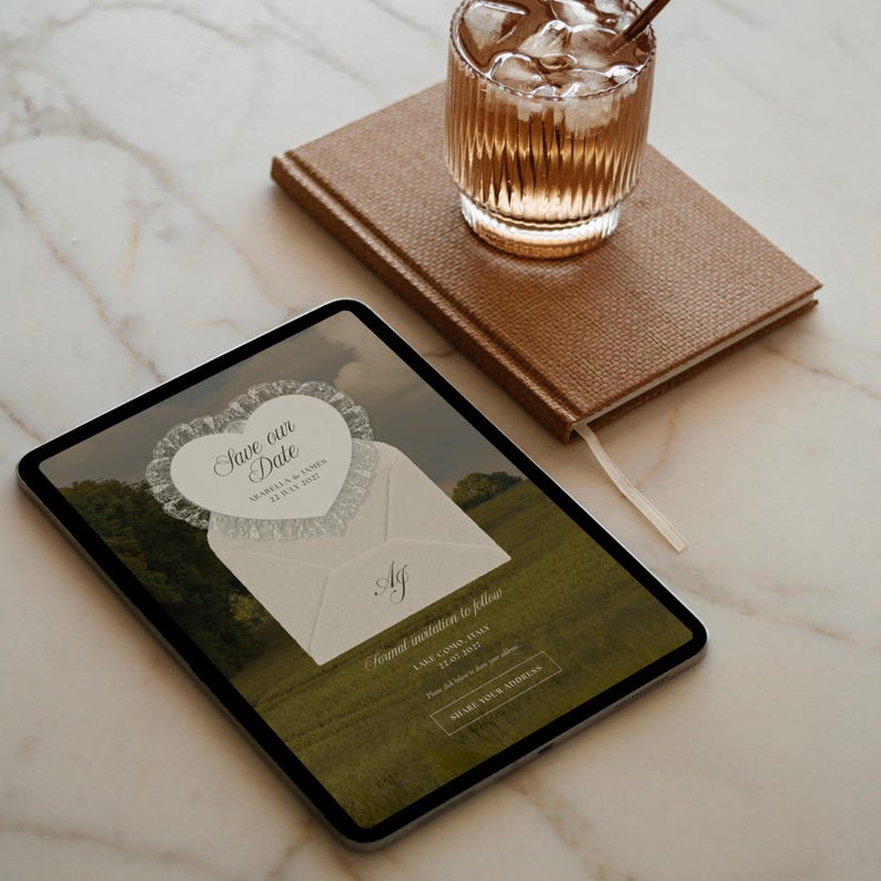 May include: A digital tablet displays a wedding invitation with the text "Save our Date" in a heart-shaped design. A brown leather-bound notebook and a glass of iced beverage sit nearby on a marble surface. The invitation includes the initials "AJ".