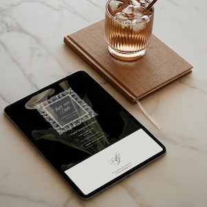 Puede incluir: Una tableta digital que muestra una invitaci&oacute;n de boda con el texto "Save our Date" en un marco decorativo, sobre un fondo negro con acentos florales. Tambi&eacute;n hay un cuaderno marr&oacute;n y un vaso de bebida helada.