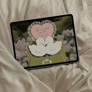 Puede incluir: Una tableta digital que muestra un dise&ntilde;o de invitaci&oacute;n de boda con un coraz&oacute;n rosa con el texto We're getting married TUSCANY, ITALY 09.09.2027, dos cisnes blancos recortados con los nombres Cecilia y Oliver, y un sobre verde con un monograma blanco.