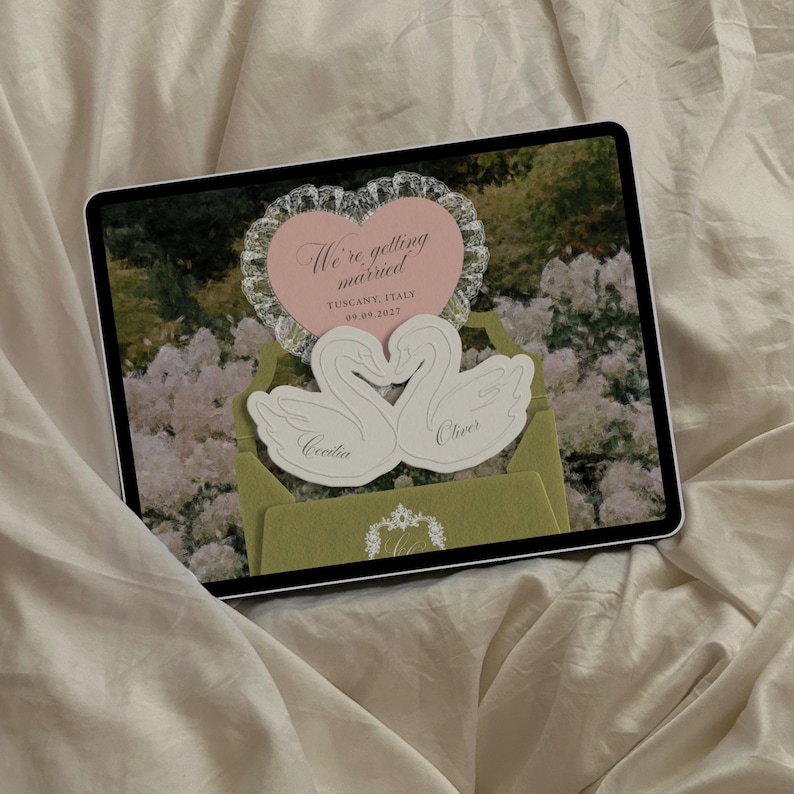 Puede incluir: Una tableta digital que muestra un dise&ntilde;o de invitaci&oacute;n de boda con un coraz&oacute;n rosa con el texto We're getting married TUSCANY, ITALY 09.09.2027, dos cisnes blancos recortados con los nombres Cecilia y Oliver, y un sobre verde con un monograma blanco.
