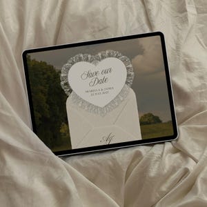 May include: A digital tablet displays a wedding invitation design. The invitation features a heart-shaped card with lace trim, reading "Save our Date" above the names Arabella & James, and the date July 22, 2027. The design is set against a landscape backdrop.