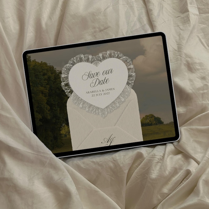 May include: A digital tablet displays a wedding invitation design. The invitation features a heart-shaped card with lace trim, reading "Save our Date" above the names Arabella & James, and the date July 22, 2027. The design is set against a landscape backdrop.