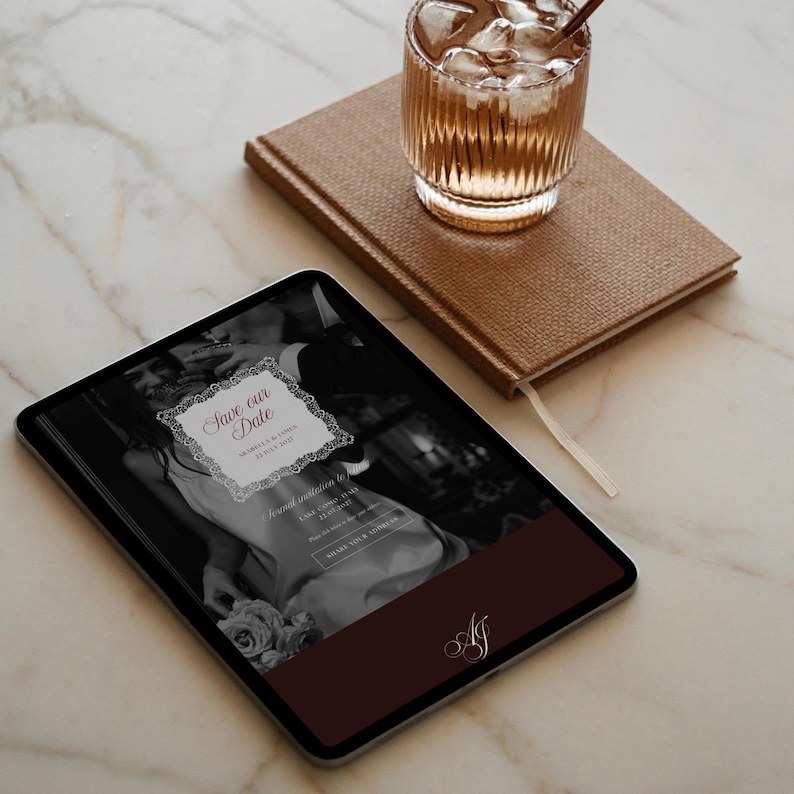 May include: A digital tablet displays a wedding invitation with the text "Save our Date" in a decorative frame. A brown leather-bound notebook and a glass of iced beverage sit nearby. The scene is set on a marble surface.