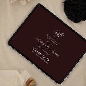 May include: A digital tablet displays a burgundy background with white elegant script. The text reads "Arabella & James" with a countdown timer. The words "We hope you can join us" are also visible. The tablet has a black border.