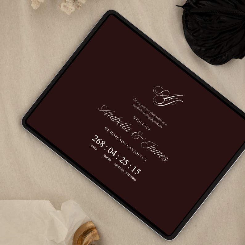 May include: A digital tablet displays a burgundy background with white elegant script. The text reads "Arabella & James" with a countdown timer. The words "We hope you can join us" are also visible. The tablet has a black border.