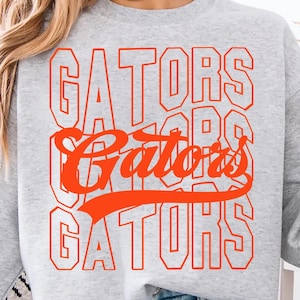 May include: A light gray sweatshirt with the word "GATORS" in orange block letters repeated three times. The word "Gators" is also written in a cursive font in the center. A person is holding a coffee mug.