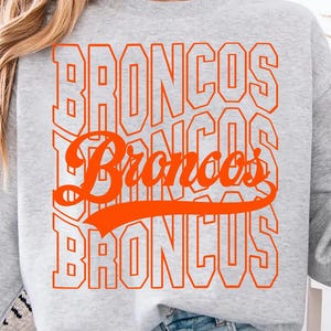 May include: A light gray sweatshirt with the word "BRONCOS" in orange block letters repeated vertically. The word "Broncos" is also written in a cursive font. A coffee mug is held in the hand.