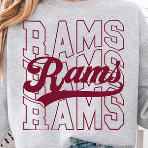 Könnte beinhalten: Ein hellgraues Sweatshirt mit dem Wort "RAMS" in einem gestapelten Design. Das mittlere Wort ist in Schreibschrift, die anderen in Blockbuchstaben. Der Text ist weinrot. Eine Kaffeetasse wird in der rechten Hand gehalten.