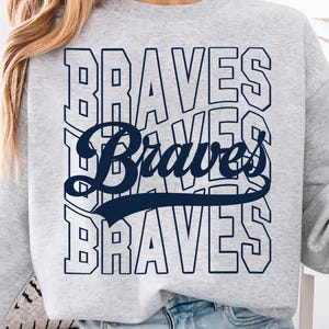 Op de afbeelding: Een lichtgrijs sweatshirt met het woord "Braves" in marineblauw. Het woord is drie keer gestapeld, met een cursief "Braves" in het midden. De sweatshirt heeft lange mouwen en een ronde hals.