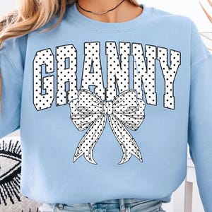 Könnte beinhalten: Hellblaues Sweatshirt mit dem Wort "GRANNY" in weißen, gepunkteten Buchstaben. Eine große weiße Schleife mit schwarzen Punkten befindet sich darunter. Das Sweatshirt hat lange Ärmel und einen Rundhalsausschnitt.