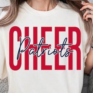 May include: A cream-colored short-sleeve t-shirt featuring the word CHEER in large, bold, red capital letters. Overlaid across the center of the red text is the word Patriots written in a dark blue, cursive script font with a decorative flourish.