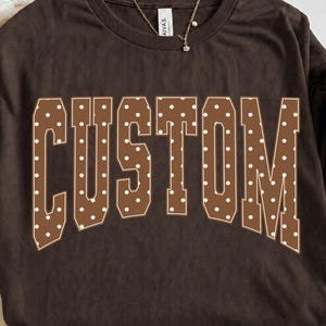 Könnte beinhalten: Ein dunkelbraunes Kurzarm-T-Shirt mit dem Wort CUSTOM, das in einer großen, geschwungenen Schriftart im College-Stil gedruckt ist. Die Buchstaben sind braun mit einem weißen Punktemuster. Eine dünne goldfarbene Kette liegt am Halsausschnitt.