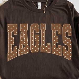 Peut inclure: Un t-shirt à manches courtes marron foncé avec le mot EAGLES en grandes lettres arquées de style bloc. Les lettres sont marron avec un motif à pois blancs et un fin contour blanc. Un collier à chaîne dorée repose au niveau de l'encolure.