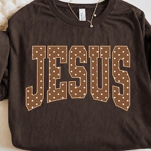 Könnte beinhalten: Ein dunkelbraunes Kurzarm-T-Shirt mit dem Wort JESUS in großen Blockbuchstaben. Die Buchstaben sind braun mit einem weißen Punktemuster und einer dünnen weißen Umrandung und befinden sich mittig auf der Brust des Kleidungsstücks.