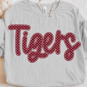 May include: A light grey short-sleeved t-shirt featuring the word Tigers in a dark red cursive font. The lettering is filled with a pattern of small white polka dots. The shirt has a crew neckline and a tag visible at the inner collar area.