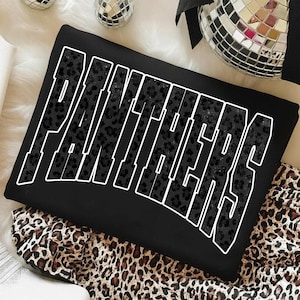 May include: Black sweatshirt with the word "PANTHERS" in large, white outlined letters. The letters are filled with a dark leopard print pattern. The sweatshirt is folded on top of a leopard print fabric.