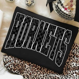 May include: Black sweatshirt with the word "HORNETS" in large white outlined letters. The letters are filled with a checkered and leopard print design. The sweatshirt is folded on a leopard print fabric.